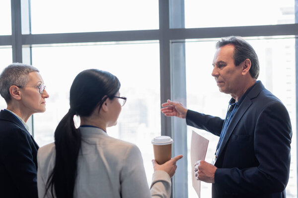 Businessman with paper folder pointing with hand near colleagues in office 