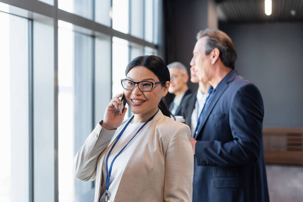 Asian businesswoman talking on smartphone near blurred colleagues 