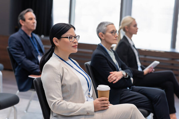 Asian businesswoman with coffee to go sitting near colleagues in conference room