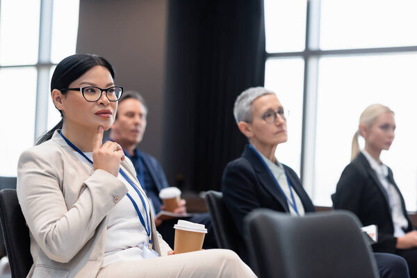 Asian businesswoman with coffee to go sitting in conference room near colleagues 