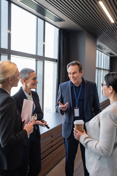 Smiling businessman pointing with hand near interracial businesswomen with smartphone and coffee to go 