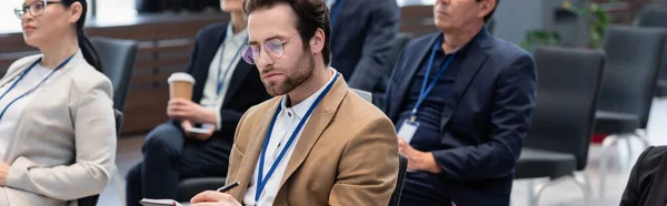 Junger Geschäftsmann Schreibt Auf Notizbuch Neben Verschwommenen Interrassischen Kollegen Konferenzraum — Stockfoto