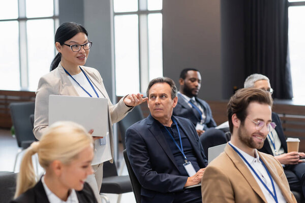 Улыбающаяся азиатская бизнесвумен с ноутбуком разговаривает рядом с деловыми людьми во время семинара 