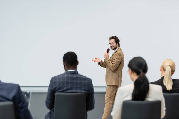 Smiling speaker talking and pointing with hand near blurred multiethnic business people during seminar 