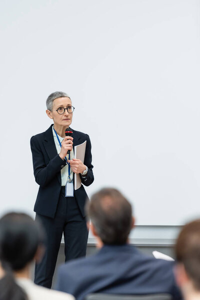 Businesswoman with paper folder and microphone talking during seminar 