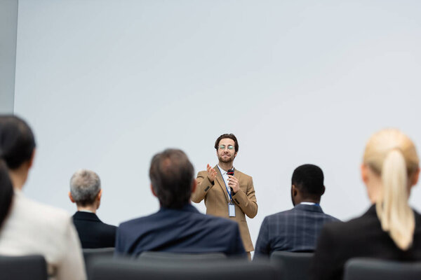 Улыбающийся оратор с микрофоном, указывающий рукой на мультиэтнических бизнесменов