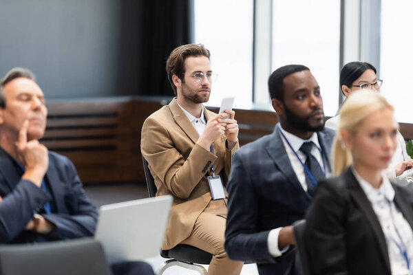 Businessman in eyeglasses using smartphone near multiethnic colleagues during seminar 