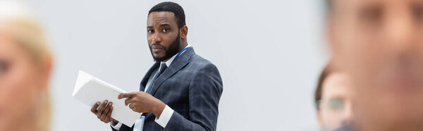 African american businessman with documents looking at camera near blurred colleagues, banner 