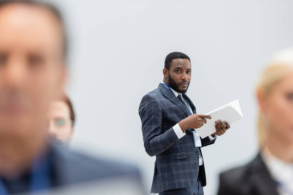 African american businessman with documents standing near blurred business people 