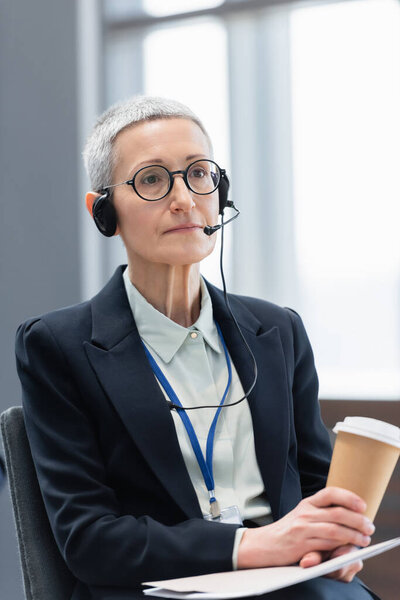 Mature businesswoman in headset holding papers and coffee to go during conference 