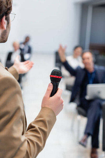 young lecturer with microphone pointing at blurred audience during seminar