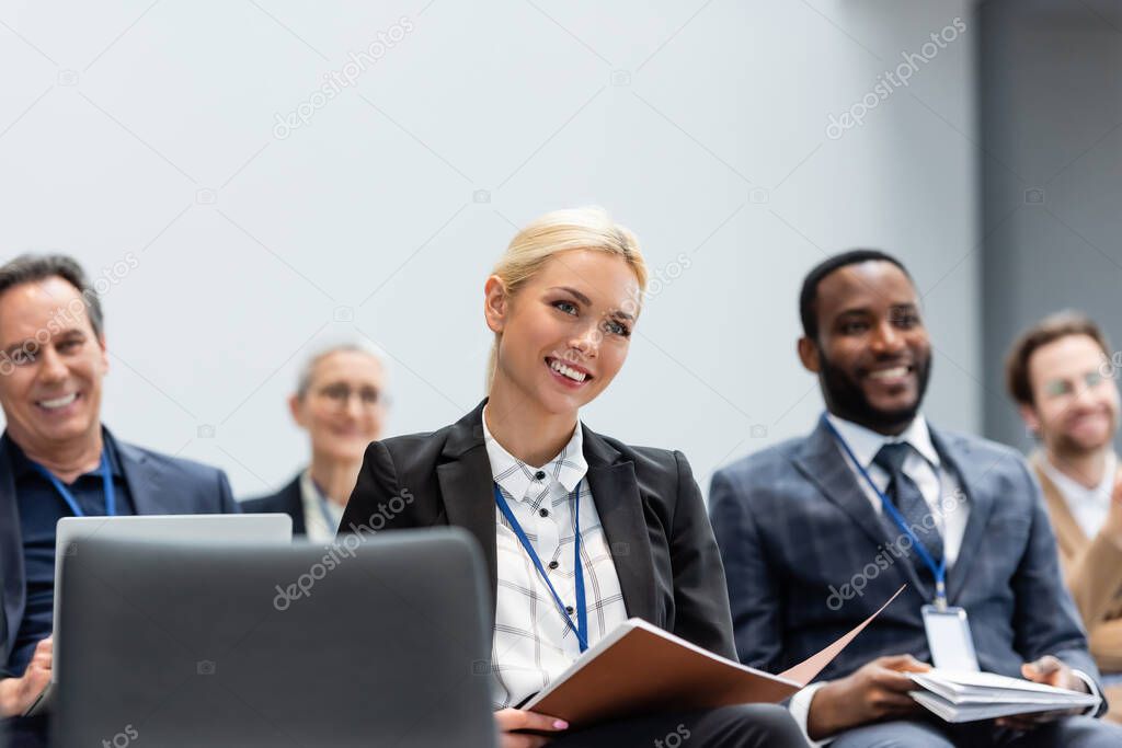 Cheerful businesswoman with paper folder sitting near multiethnic colleagues during seminar 