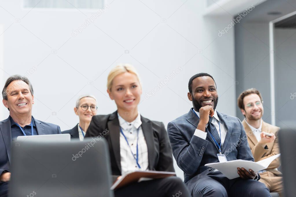 Mature businessman with laptop smiling near multiethnic colleagues in conference room 