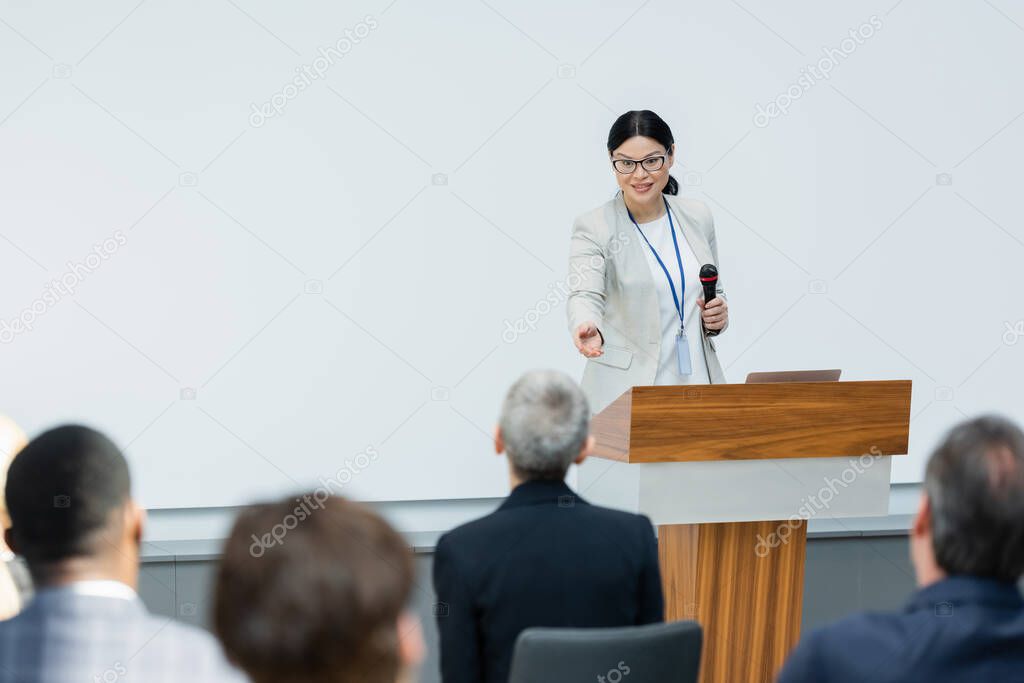 asian lecturer pointing with hand at businesswoman during seminar, blurred foreground
