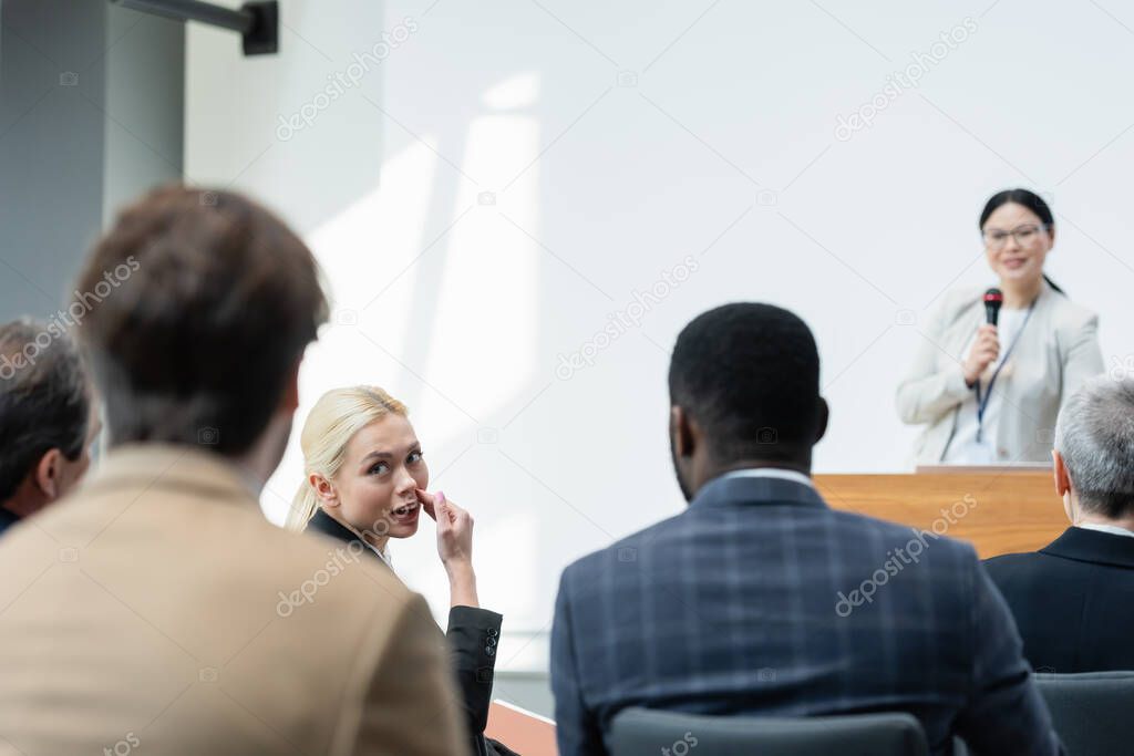 young woman talking to colleagues during seminar near asian lecturer on blurred background