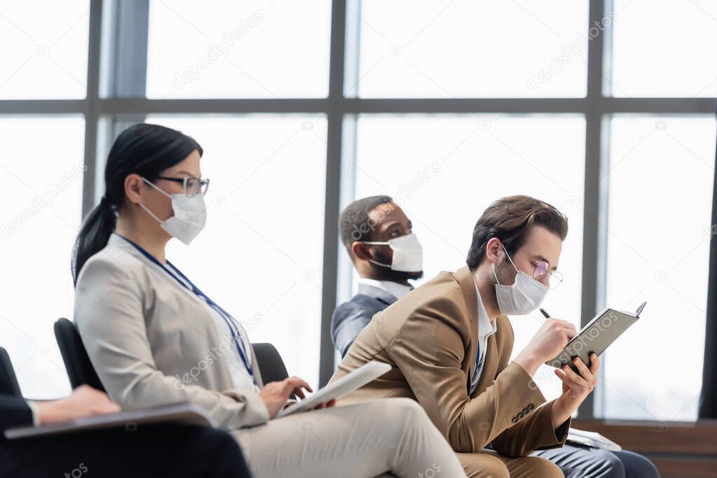 young businessman in medical mask writing in notebook near interracial colleagues