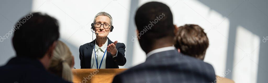 middle aged speaker pointing at blurred business people during seminar, banner