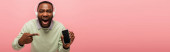 Africký Američan ve sluchátkách ukazuje na smartphone s prázdnou obrazovkou izolované na růžové, banner 
