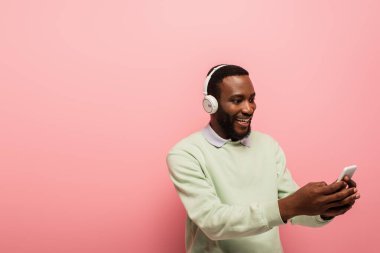 Gülümseyen Afro-Amerikan kulaklıklı adam pembe arka planda akıllı telefon kullanıyor. 