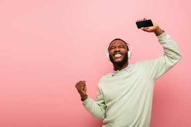 Kulaklıklı Afro-Amerikan bir adam akıllı telefon tutuyor ve evet işareti yapıyor. Pembe üzerine izole edilmiş. 