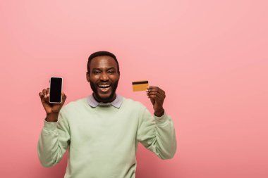 Neşeli Afrikalı Amerikalı adam elinde cep telefonu ve pembe arka planda kredi kartı tutuyor. 