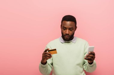 Pembe arka planda online alışveriş yaparken kredi kartı ve akıllı telefon kullanan Afrikalı Amerikalı bir adam. 