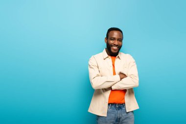 Pozitif Afro-Amerikalı adam kameraya gülümserken kollarını çapraz bağlamış mavinin üzerinde tek başına duruyordu.
