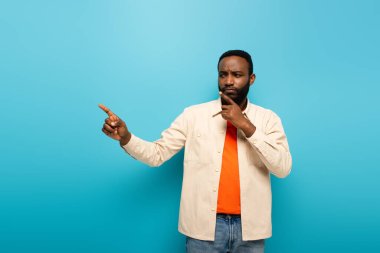 thoughtful african american man touching face while pointing aside with finger on blue background clipart