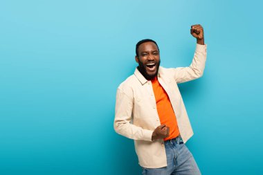 Heyecanlı Afrikalı Amerikalı adam mavi arka planda zafer jesti gösterirken kameraya bakıyor.