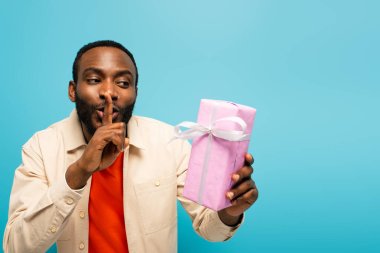 young african american man showing secret sign while holding gift box isolated on pink clipart
