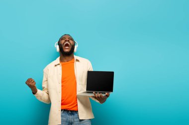 Heyecanlı Afro-Amerikan kulaklıklı adam mavi arka planda dizüstü bilgisayarını tutarken sevinç jesti yapıyor.