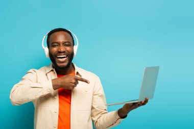 Kulaklıklı gülen Afro-Amerikan adam mavi arka planda dizüstü bilgisayarı işaret ediyor.