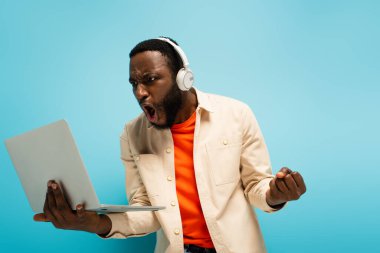 Kızgın Afro-Amerikan kulaklıklı adam mavi dizüstü bilgisayara bakarken yumruğunu sıkıyor.