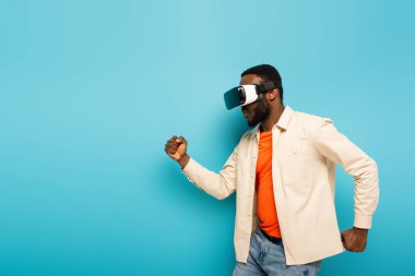 Afro-Amerikan adam mavi arka planda VR kulaklıkla dans ediyor.
