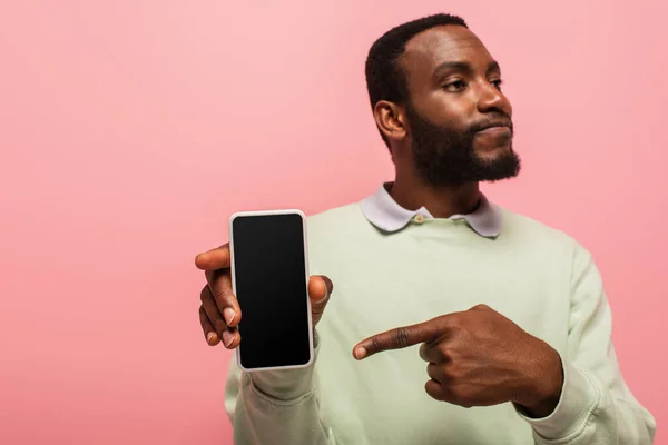 Blurred african american man pointing at smartphone with blank screen isolated on pink