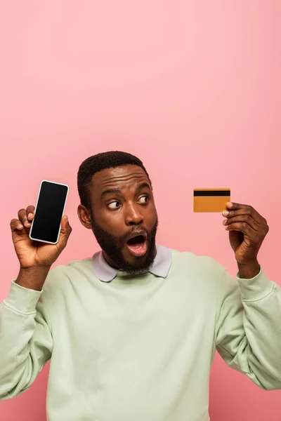 Zszokowany Afrykański Amerykanin Patrząc Kartę Kredytową Trzymając Smartfon Odizolowany Różowy — Zdjęcie stockowe