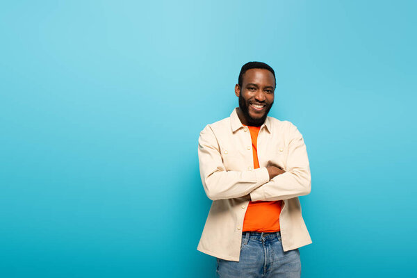 positive african american man smiling at camera while standing with crossed arms isolated on blue