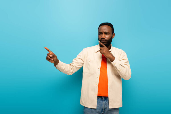 thoughtful african american man touching face while pointing aside with finger on blue background