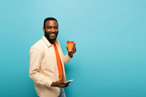 Улыбающийся Африканский Американец Держащий Выпивку Вынос Используя Мобильник Изолированный Голубом — стоковое фото