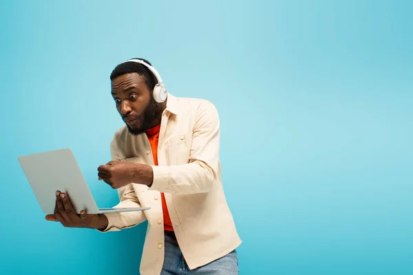 angry african american man in headphones showing clenched fist near laptop on blue background