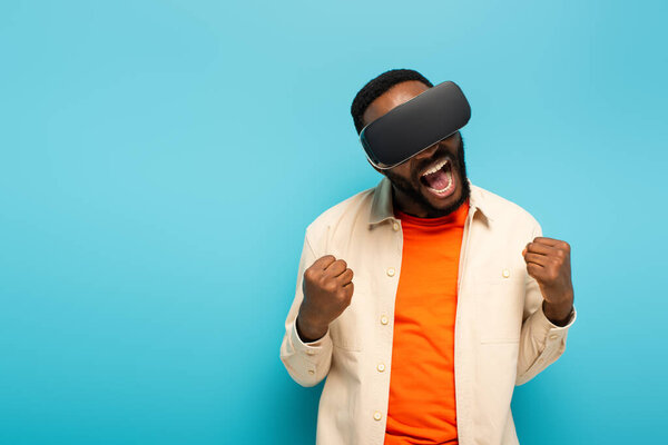 excited african american man in vr headset showing rejoice gesture isolated on blue