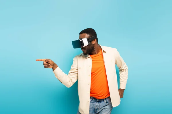 young african american man pointing aside with finger while gaming in vr headset on blue background