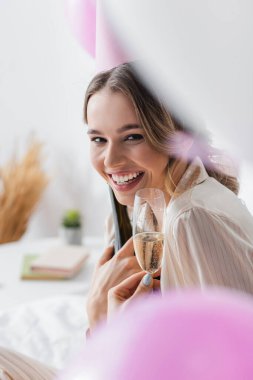 Parti şapkalı gülümseyen kadın bulanık balonların yanında şampanya kadehi tutuyor. 