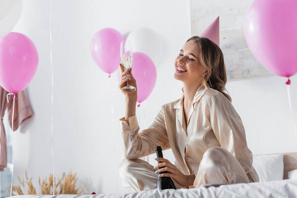 Cheerful woman with champagne celebrating birthday near balloons in bedroom 