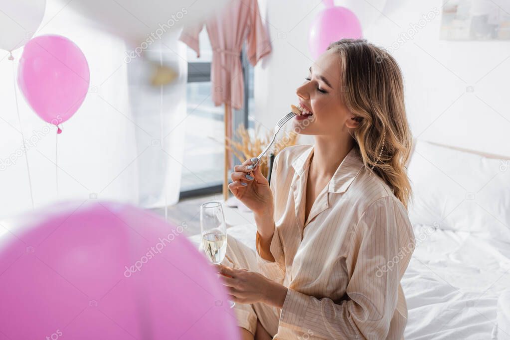 Smiling woman with champagne earing banana near balloons in bedroom 