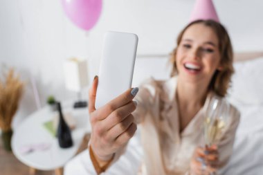 Bulanık bir kadının elinde akıllı telefon ve yatak odasında şampanya ile doğum günü kutlaması. 