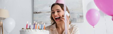 Balonların ve doğum günü pastasının yanında cep telefonuyla konuşan heyecanlı bir kadın. 