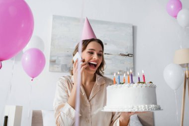 Doğum günü pastalı kadın akıllı telefondan konuşuyor ve evde balonların yanında doğum günü pastası tutuyor. 