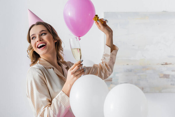 Smiling woman with party horn and champagne celebrating birthday near balloons at home 