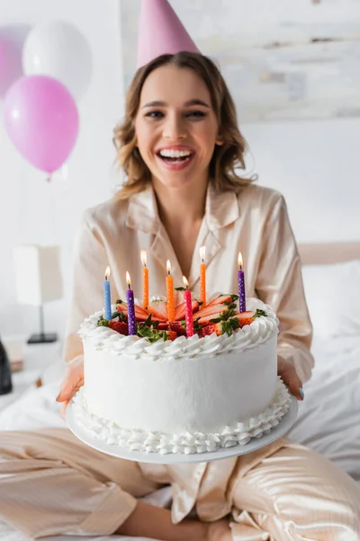Birthday cake with candles in hands of blurred woman on bed